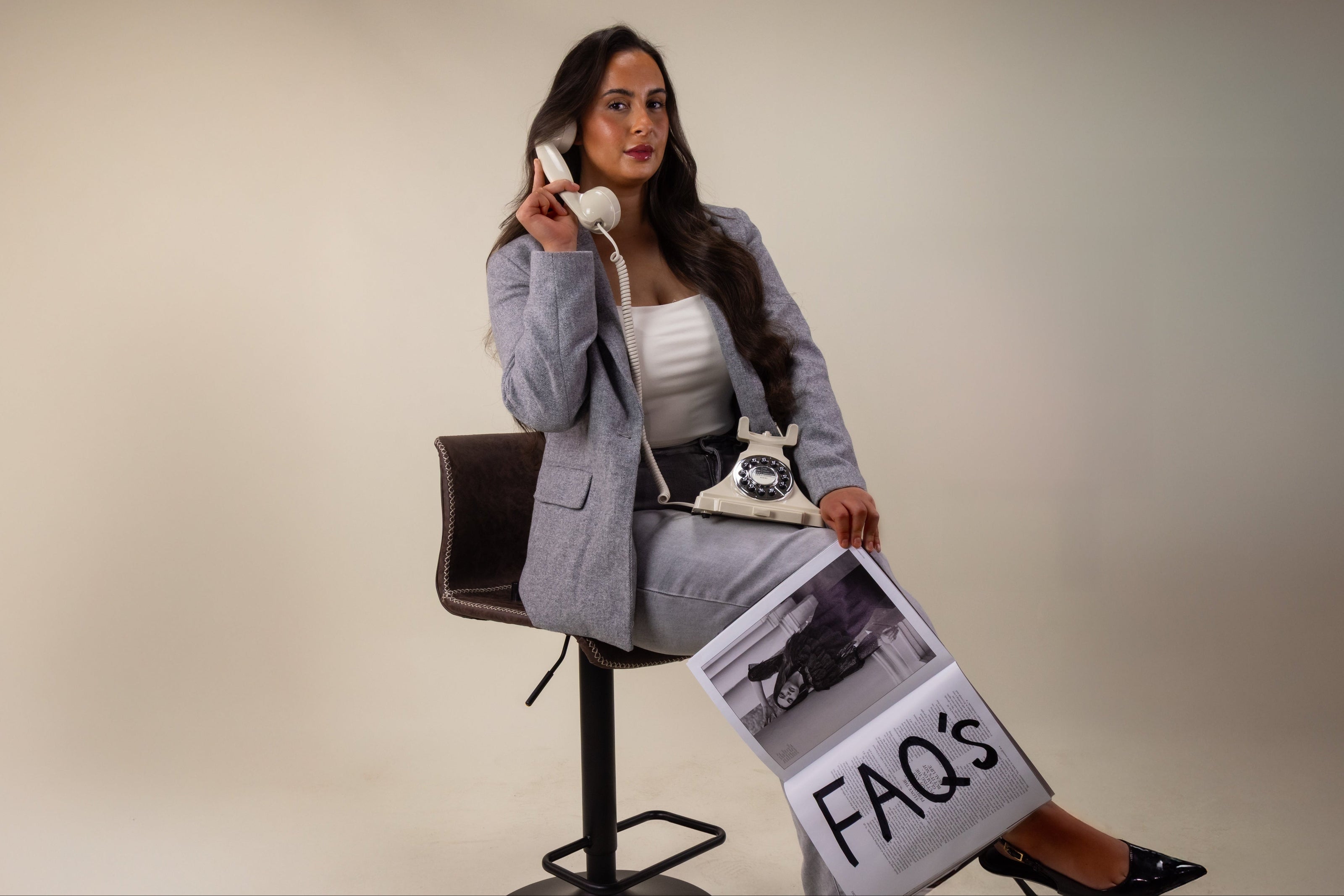 Woman sitting on a stool holding a phone and a newspaper with 'FAQs' on a plain background